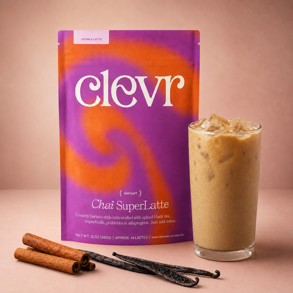 A pouch of Clevr Chat SuperLatte instant drink mix stands next to a tall glass of iced latte on a warm beige background, with cinnamon sticks and vanilla beans placed in front.