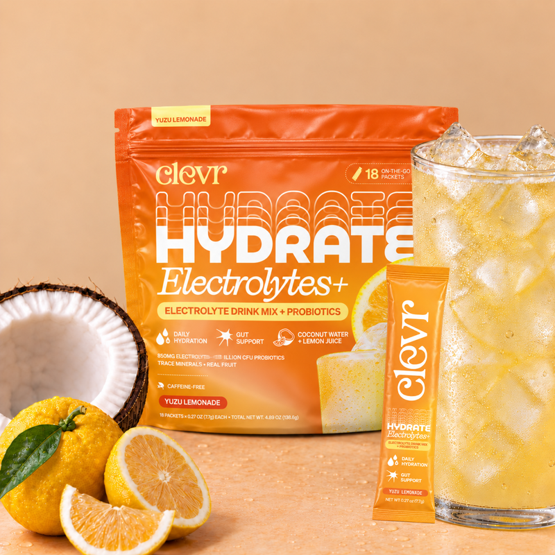 Clevr Hydrate Electrolytes+ pouch in Yuzu Lemonade flavor, shown with a single stick packet, a glass of iced yellow drink, and lemon/coconut props on a warm beige background.