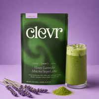 A package of Clevr Honey Lavender Matcha SuperLatte stands next to a glass of green matcha latte on a purple background, with lavender flowers and a small pile of matcha powder in front.