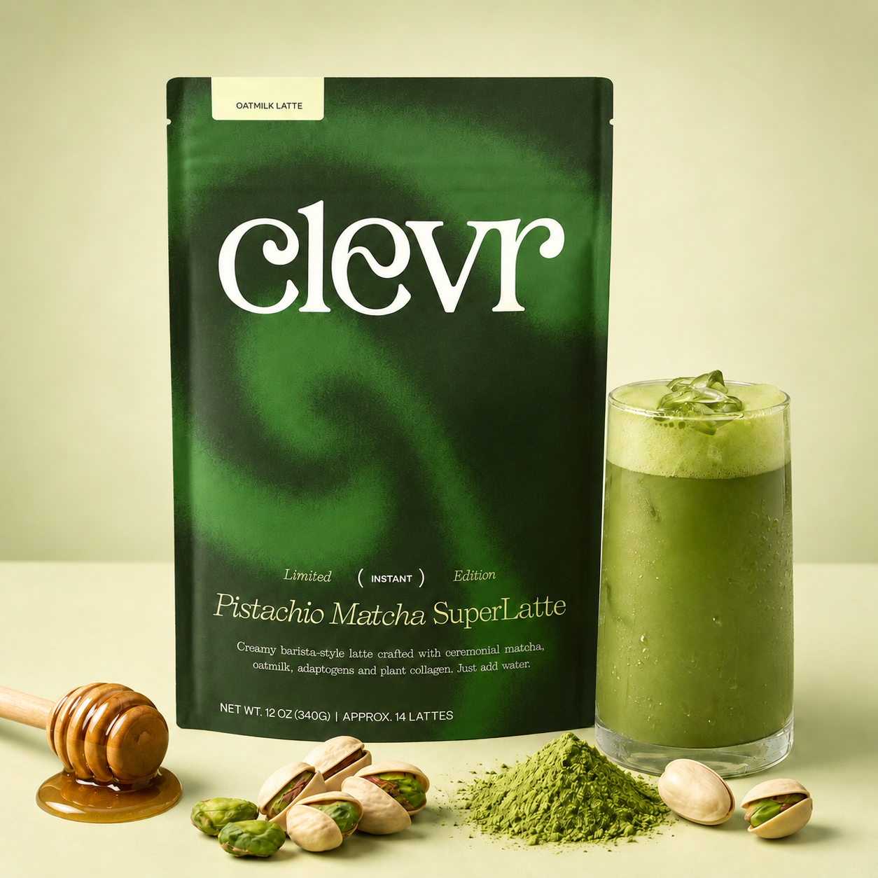 A package of Clevr Pistachio Matcha SuperLatte stands on a light beige surface beside a glass of green matcha latte, with pistachios, a honey dipper, and a small pile of matcha powder in front.
