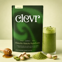 A package of Clevr Pistachio Matcha SuperLatte stands on a light beige surface beside a glass of green matcha latte, with pistachios, a honey dipper, and a small pile of matcha powder in front.
