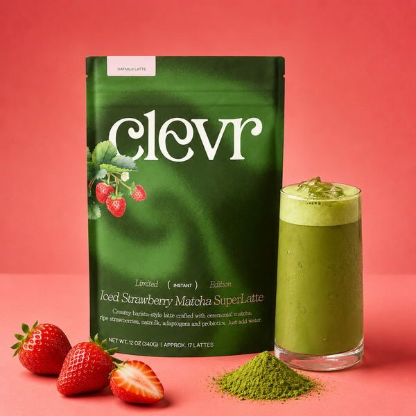 A bag of Clevr Iced Strawberry Matcha SuperLatte stands on a pink studio background next to a tall glass of green matcha latte and a small pile of matcha powder, with whole and sliced strawberries in front.